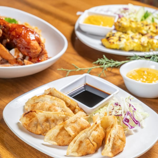 A close-up of golden-fried Thai dumplings with soy sauce, coleslaw, and other dishes in the background.